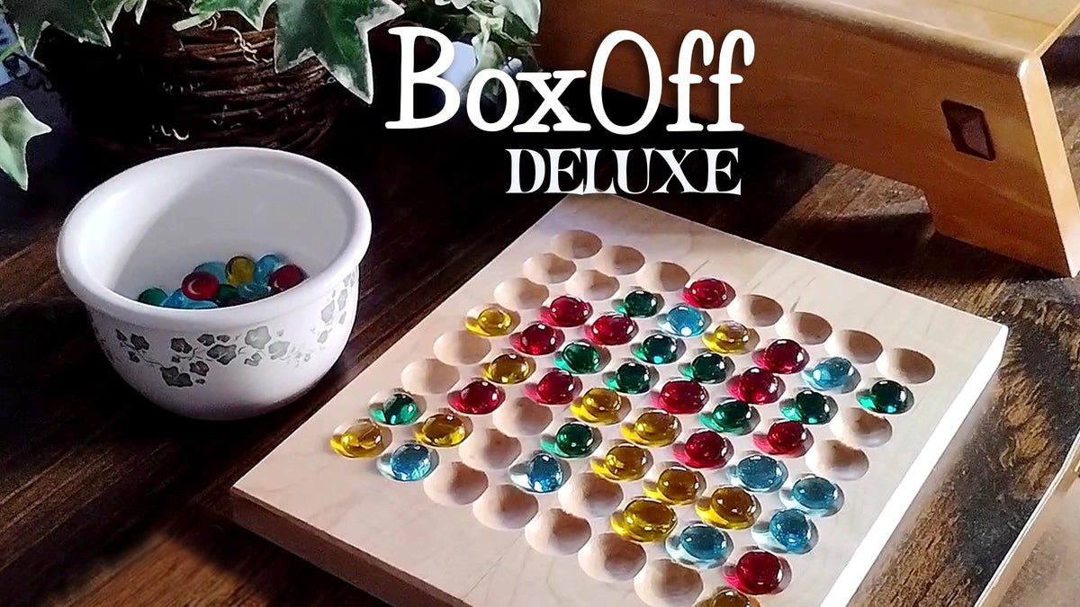 A wooden game board with many colorful round gems placed in 64 indentations, shown on a dark wooden table next to a white bowl of additional gems. Large 'BoxOff DELUXE' text is overlaid at the top.