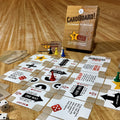 The 'Cardboard!' board game set arranged on a wooden tabletop, featuring a brown cardboard box, various white and brown game tiles, dice, and colorful player pawns.