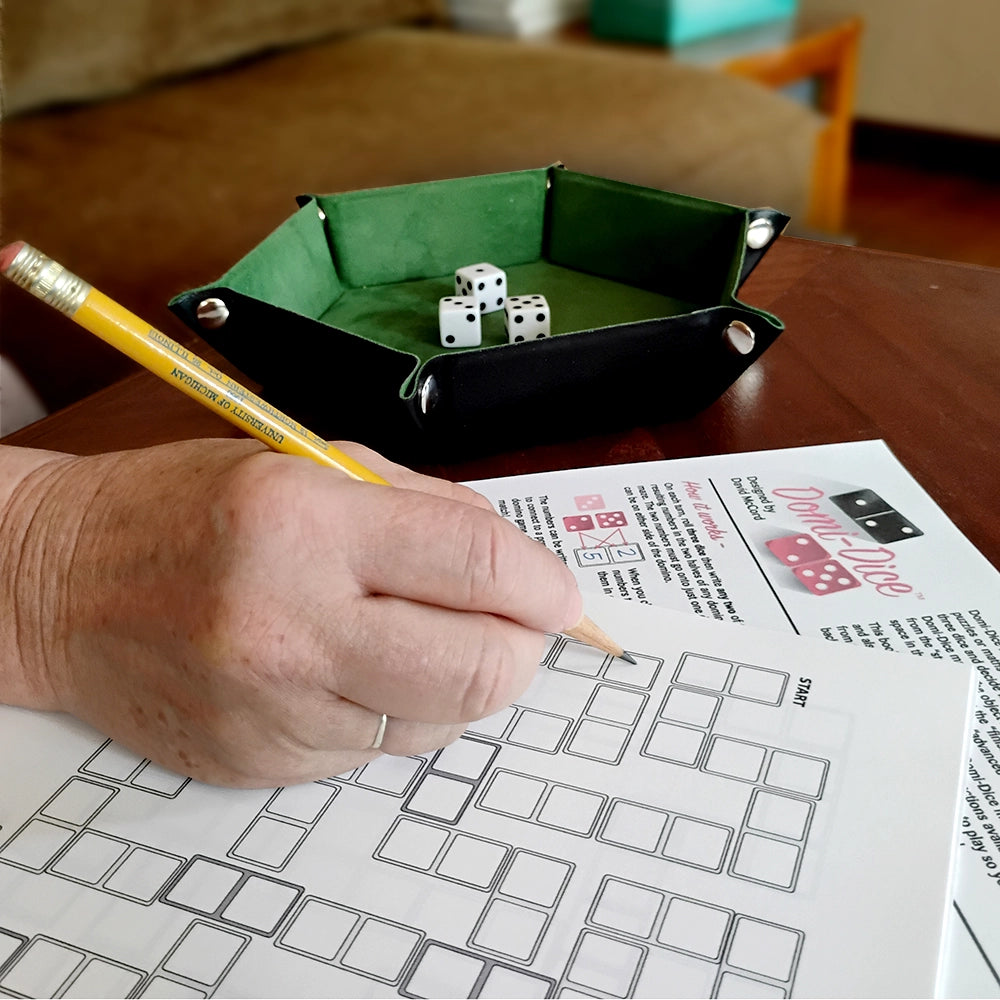 A person's hand holding a pencil and writing on a printed game sheet, with a black and green dice tray containing white dice on a wooden table in the background.
