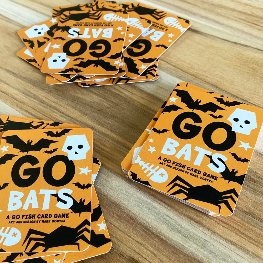 Several stacks of orange GO BATS arranged on a light wood tabletop. The cards feature black bat silhouettes, white skulls and fishbone graphics, and bold black-and-white text.