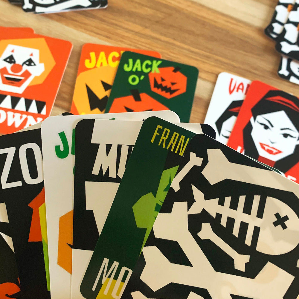 A close-up of a fanned deck of Halloween-themed cards on a wooden tabletop, with several cards partially visible in the background showing clown, jack-o'-lantern, and vampire artwork.