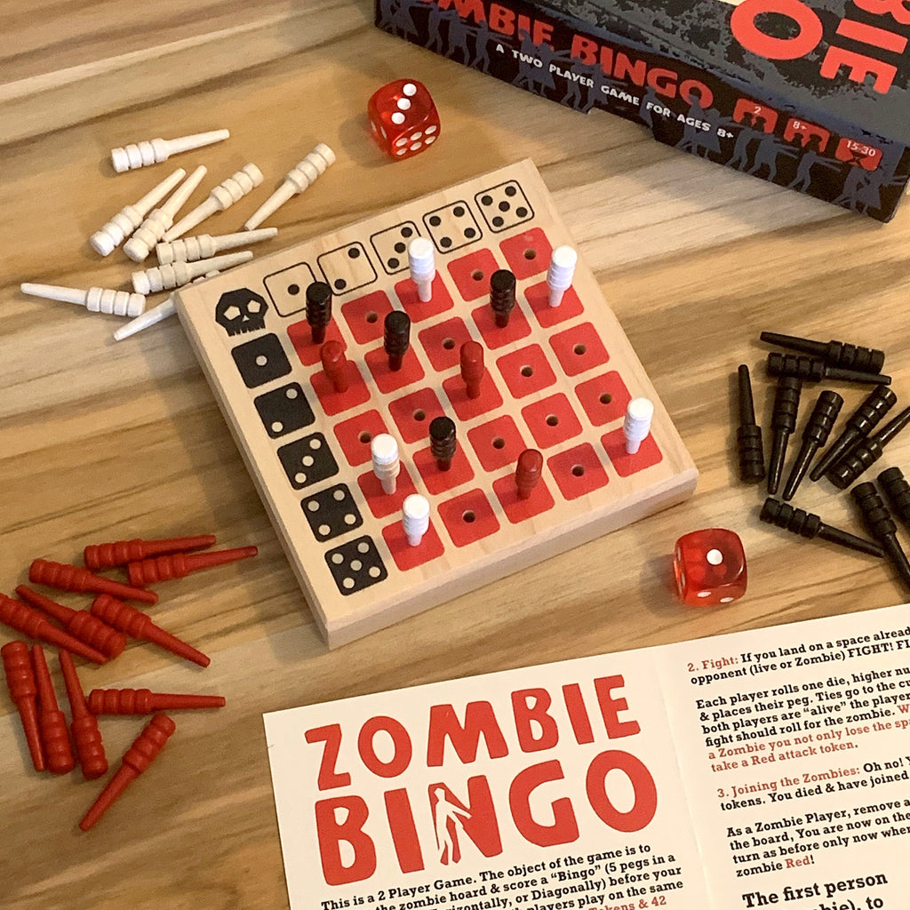Zombie Bingo Game board set on a wooden tabletop, with the game board, red and black dice, white and black zombie tokens, printed game instructions, and a black box labeled 'Zombie Bingo Game'.