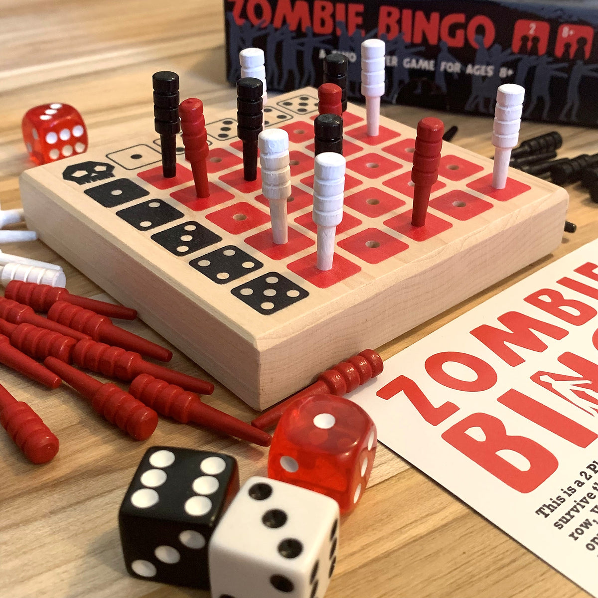 Zombie Bingo board game set on a wooden tabletop, featuring a light wood game board with red and black bingo squares, red and white bingo players, red and black dice, loose red and black tokens, and part of the product box and instruction card visible.